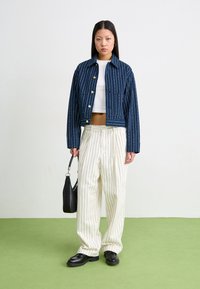 Modrá vzorovaná denimová bunda se zlatými knoflíky, doplněná o bílé kalhoty s jemným proužkem. Černá kabelka a mokasíny dotvářejí celkový outfit.
