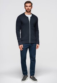 Dunkelblaues Zip-Up Sweatshirt mit Rundhalsausschnitt, gerippten Bündchen und Saum, kombiniert mit dunklen Jeans und grauen Schnürschuhen.