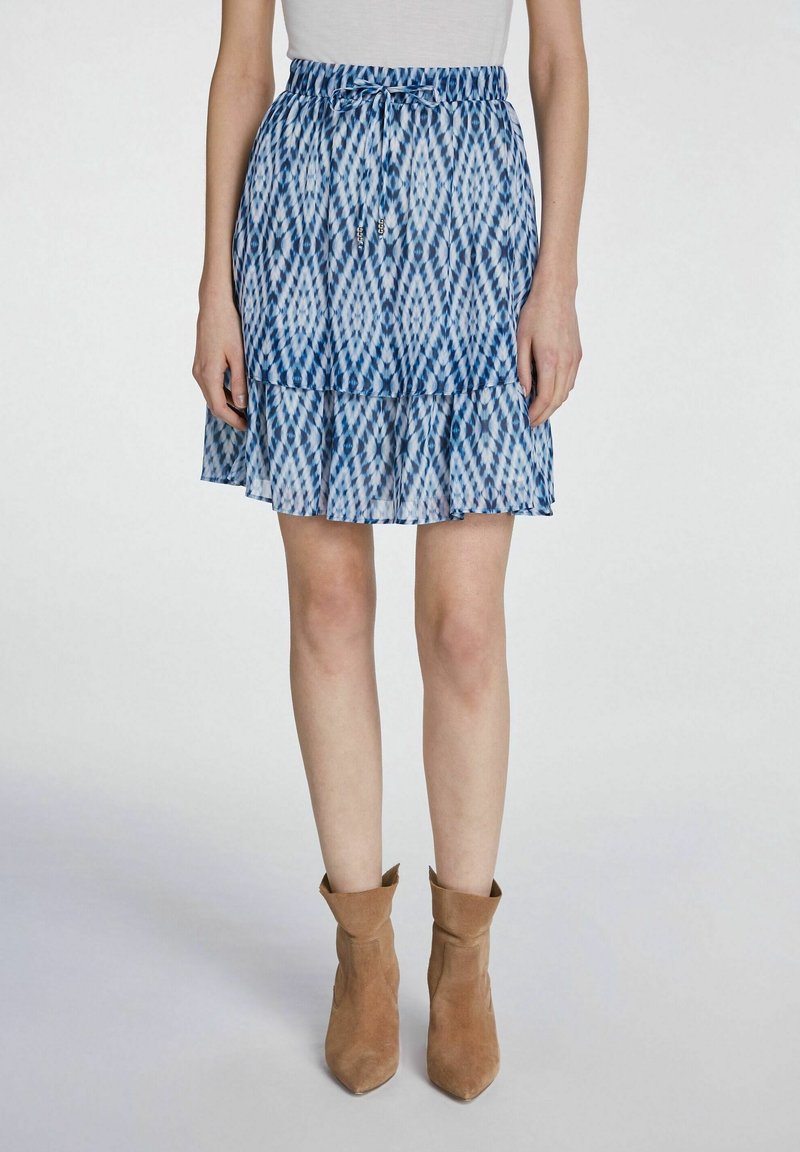 Jupe à pans bleue à motifs avec un ourlet en volants et une taille élastique. Portée avec des bottines beige, mettant en valeur un tissu léger et fluide.