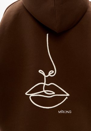Sweat à capuche marron avec un dessin en lignes blanches représentant un visage aux lèvres et au nez bien marqués. Comprend le nom de la marque "MIRONS" brodé en petit texte.