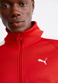 Rød zip-up jakke med ribbet krave, prydet med hvide detaljer på skuldrene og et hvidt Puma-logo på brystet. Struktureret stof.