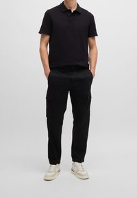 Homme portant un polo noir à manches courtes, un pantalon cargo noir avec des poches, et des baskets blanches, se tenant avec les mains dans les poches.