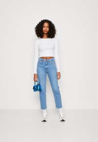 Top curto de manga longa branco, jeans de cintura alta azul-claro e sapatilhas brancas. A segurar uma pequena mala azul brilhante. Textura suave.