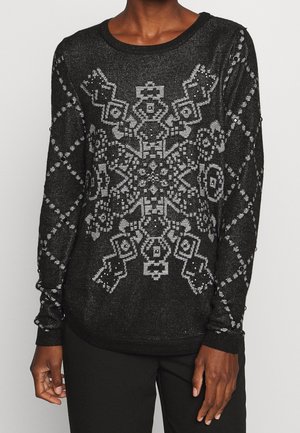Personne portant un pull noir à manches longues avec un grand motif géométrique de flocon de neige blanc et des motifs en forme de losange sur les manches.