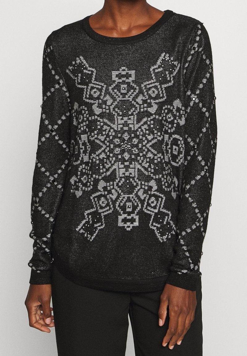 Personne portant un pull noir à manches longues avec un grand motif géométrique de flocon de neige blanc et des motifs en forme de losange sur les manches.
