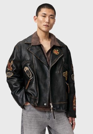 Aziatische man die een zwarte leren jas draagt met kleurrijke geborduurde patches, een bruin shirt en grijze spijkerbroek, staand tegen een effen achtergrond.