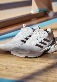 Białe buty sportowe z czarnymi akcentami, siateczkowa cholewka, teksturowany wzór, gumowa podeszwa oraz wyraźne logo z trzema paskami po bokach.