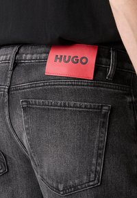 Vue arrière d'une personne portant un jean noir avec une étiquette rectangulaire rouge affichant le logo "HUGO" sur la ceinture.