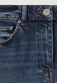 Nærbillede af blå denimjeans, der viser forlomme, bæltestrop, orange syning og sølvknap med indgraveret tekst.