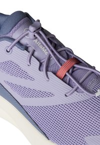 Lilla sportsko med teksturert mesh-overdel, glatte pålegg, elastiske lisser og en kontrasterende rød aksent. Gummisåle synlig.