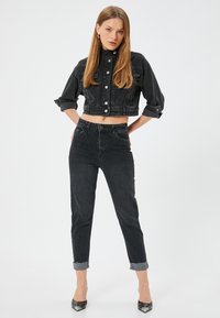 Chaqueta de mezclilla negra cortada con un cuello alto y cierre de botones, combinada con pantalones ajustados negros con los puños doblados.