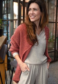 Langsleeve, open-front cardigan in textuurrijke roestrode breisteek, gedragen over een lichtgrijze top en bijpassende joggers, met zijzakken.