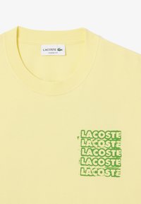 T-shirt en coton jaune avec un col rond côtelé et un imprimé logo superposé vert sur le côté gauche. Design à coupe classique.