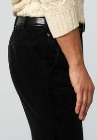 Schwarze Cordhosen mit vertikalem Rippmuster, figurbetontem Schnitt und Seitentasche. Kombiniert mit einem strukturierten, hellen Strickpullover. Bund sichtbar.