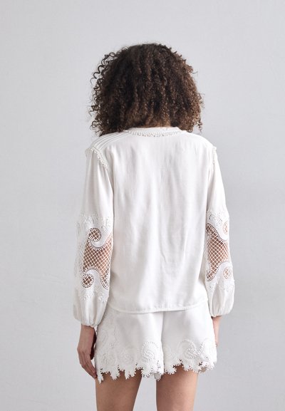 Vit blus med genomskinliga broderade spetsärmar och vågig fåll, med texturerade detaljer och en avslappnad passform.