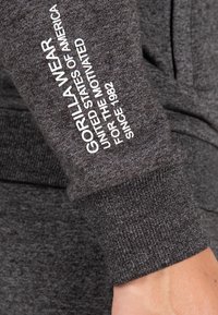 Siva dukserica od tkanine s rebrastim manžetama. Sadrži bijeli natpis "GORILLA WEAR UNITED STATES OF AMERICA FOR THE MOTIVATED SINCE 1982."