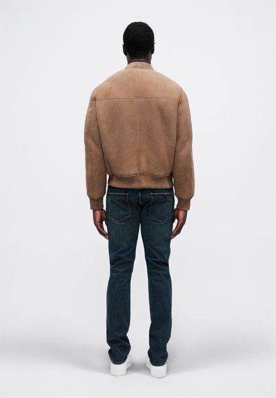 Férfi barna velúr dzsekit, sötétkék farmernadrágot és fehér sportcipőt viselve, háttal áll egy sima világos háttér előtt.