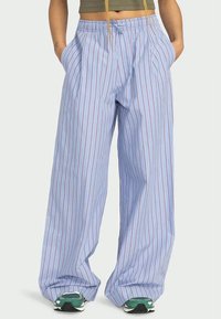 Pessoa vestindo calças largas, de um azul claro, com finas riscas verticais vermelhas, uma blusa curta em verde-oliva, ténis verdes, com as mãos nos bolsos.
