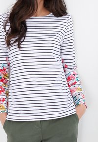 Randig vit och marinblå långärmad topp med blommönstrade accenter på ärmarna, kombinerad med gröna byxor. Mjuk tyg, avslappnad passform.