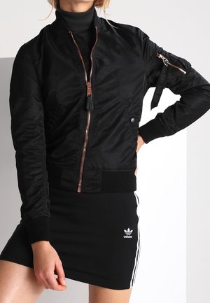 Personne portant une veste bomber noire avec une fermeture éclair à l'avant et une mini-jupe Adidas noire avec des rayures blanches et un logo.