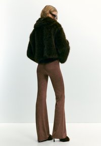 Donna che indossa una giacca pelosa marrone scuro, pantaloni a campana marroni abbinati, pantofole pelose con tacco alto e grandi occhiali da sole tartarugati, voltata di spalle.