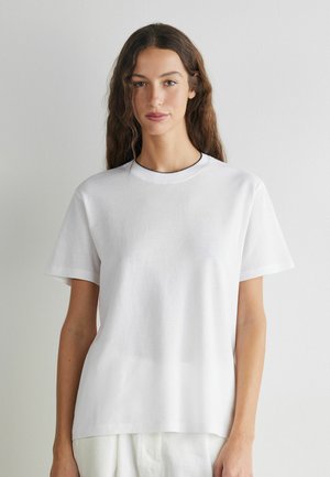 Frau mit langen braunen Haaren, die ein schlichtes weißes T-Shirt mit kurzen Ärmeln trägt und vor einer hellgrauen Wand steht.