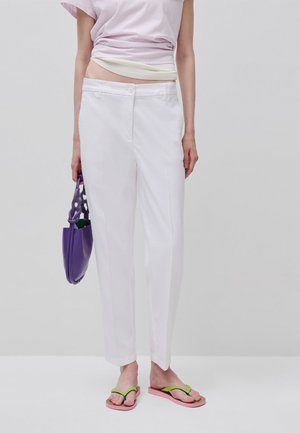 Person trägt weiße maßgeschneiderte Hose, hellrosa Oberteil, hält eine violette Handtasche und trägt pink-grüne Flip-Flops vor einfarbigem Hintergrund.