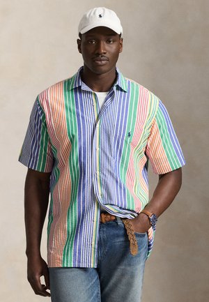 Mand iført en flerfarvet kortærmet skjorte med lodrette striber, hvid kasket med navy logo, blå jeans, flettet bælte og ur, der poserer mod en ensfarvet baggrund.