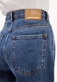 Jean en denim bleu avec des coutures orange, doté d'une étiquette en cuir portant la mention "ARMEDANGELS" au dos de la taille. Comprend deux poches arrière.