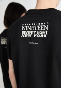 Camiseta de algodón negra con texto blanco en la parte trasera: "ESTABLECIDO EN MIL NOVECIENTOS SETENTA Y OCHO NUEVA YORK" y el logo de "Calvin Klein Jeans".
