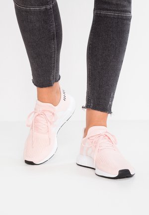 Persoon die lichtroze sportieve sneakers draagt met witte zolen en zwarte accenten, gecombineerd met zwarte cropped skinny jeans.