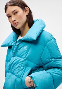 Tirkizna puffer jakna s velikim ovratnikom, kvadratnim dizajnom i srebrnim detaljima. Ima logo na prsima i teksturiranu površinu.