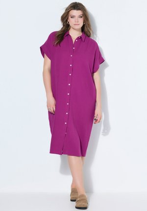 Kvinde iført løs, kortærmet, knappet magentarød kjole og beige slip-on sko, der står mod en hvid baggrund.