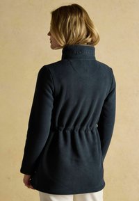 Námořnická fleecová bunda s vyvýšeným límcem, staženým pasem a vyšitým názvem značky na límci. Měkká textura a hladký povrch.