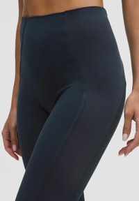 Közelkép egy személyről, aki sötét magas derékvonalú sportleggingset visel, látható oldalvarrásokkal, és a kezét lazán az oldaluknál tartja.