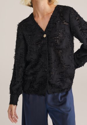 Femme portant un chemisier noir texturé à manches longues avec un bouton doré et un pantalon large en satin bleu marine sur un fond uni.