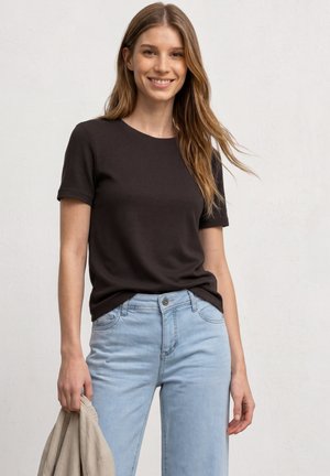 Femme souriante aux longs cheveux châtain clair portant un t-shirt noir à manches courtes et un jean bleu clair, tenant une veste beige dans sa main gauche.