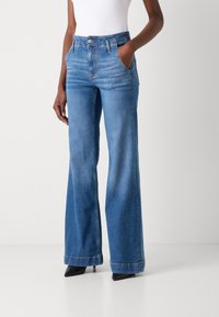 Jean bleu à jambe large avec une taille haute, fabriqué en denim. Présente un style traditionnel à cinq poches et une couture robuste. Bas droit aux chevilles.