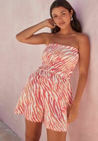 Jumpsuit bez naramenica u crvenoj i bijeloj zebraste uzorke, s smockiranim korzetom i širokim kratkim hlačama. Izrađen od laganog, teksturiranog materijala.