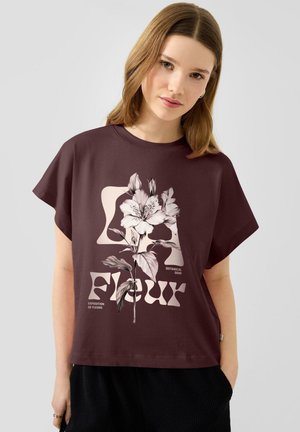 Jeune femme portant un t-shirt marron foncé avec un grand motif floral rose clair et du texte, debout devant un fond clair uni.