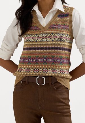 Frau trägt einen gemusterten, gestrickten ärmellosen Pullover über einem weißen Hemd mit Knöpfen, kombiniert mit brauner Hose und einem dunklen Gürtel mit silberner Schnalle.