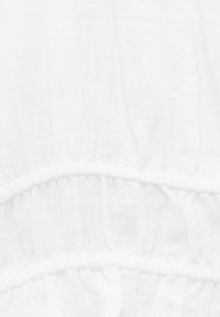 Hvidt tøj lavet af letvægts, tekstureret stof med flæser langs sømmene. Materialet ser gennemsigtigt og luftigt ud.