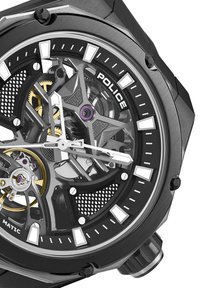 Črna kovinska ura s prozornim številčnico, vidnimi zobniki, zlato obrobo, belimi označevalci ur in drzno šestkotno obliko ohišja.