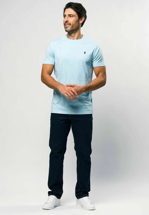 Vyras dėvi šviesiai mėlyną marškinėlius, tamsius džinsus ir baltus sportinius batelius, stovi sukryžiavęs rankas ir žiūri į šoną priešais paprastą foną.