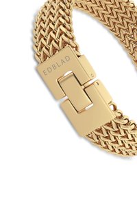 Kuldse tooniga metallist käevõru, millel on kootud disain. Omab ristkülikukujulist haaknõela, millel on graveering "EDBLAD." Sile ja läikiv pind koos omavahel põimunud lülidega.