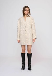 Beige patroonjas met geometrisch ontwerp, met een kraagloze halslijn en knopen, gecombineerd met hoge zwarte leren laarzen.