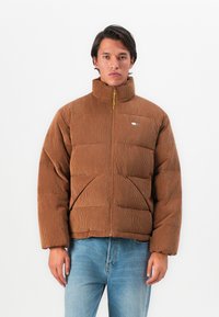 Man in een bruine corduroy pufferjack met een gele rits en een blauwe spijkerbroek, staand tegen een effen witte achtergrond.