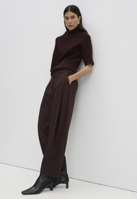 Woman leaning against a wall wearing a dark brown half-sleeve sweater, matching high-waisted wide-leg pants, and black ankle boots.