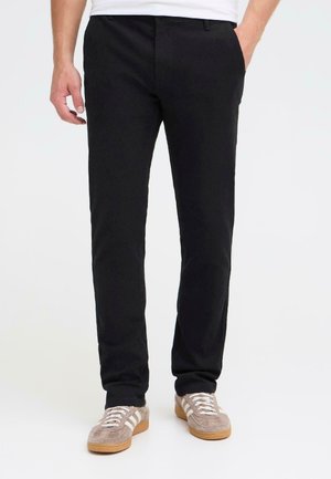Black trousers with a straight fit, made from a soft fabric. Front pockets and slightly tapered legs. Worn with light brown sneakers.