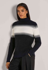 Frau mit langen dunklen Haaren, die einen schwarz-weißen gerippten Strickpullover und schwarze Hosen trägt, steht vor einer schlichten hellen Wand.